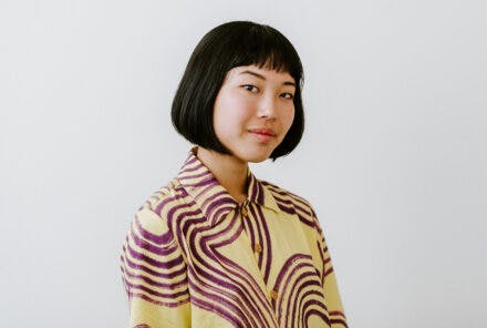 Maki smiling at the camera. She wears a yellow top with purple patterns on it.